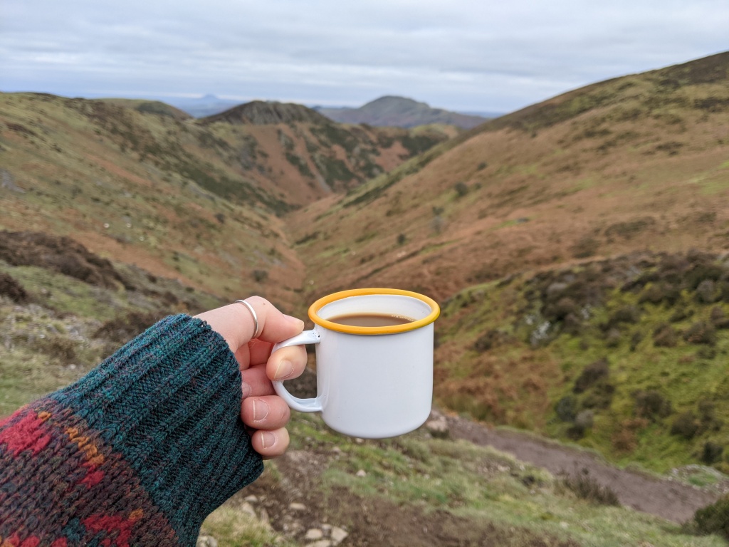 A tin mug of coffee held up against a backdrop of a rugged valley.